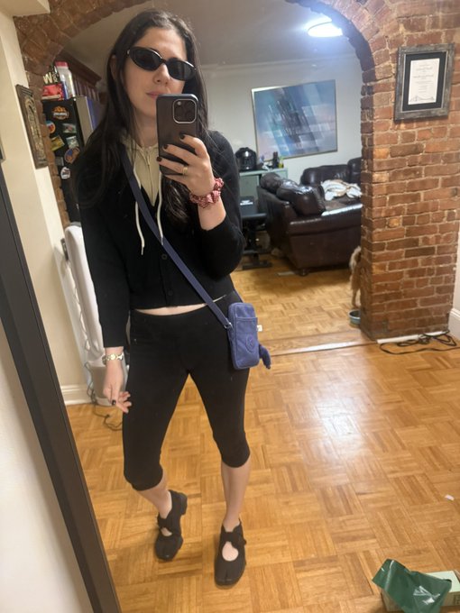 A mirror selfie of a woman wearing sunglasses, a black cropped hoodie, black capri leggings, and black ballet flats, carrying a small purple crossbody bag in a home with parquet wood floors.