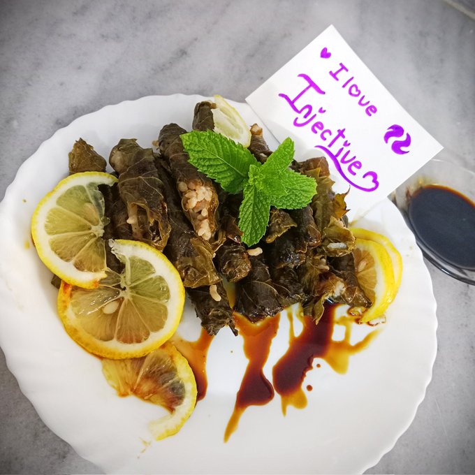 The image shows a plate of food consisting of several pieces of grape leafs, lemon wedges, and herbs, along with some sauce drizzled over them. A note is placed above the dish that reads "I love @Injective The tastiest grape leaves ever $INJ". This message emphasizes the deliciousness of the food on the plate in relation to a particular person named "@Injective".