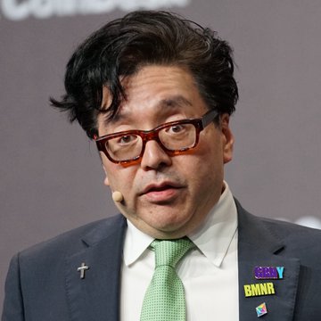 A man with dark hair, thick-rimmed glasses, and a gray suit with a green tie speaks at what appears to be a Bloomberg conference, with visible "SMNR" and "ZERO" badges on his lapel pin. The image appears somewhat unrelated to the post about Ethereum price predictions, showing what looks like a financial or business speaker rather than directly illustrating the crypto market forecast.