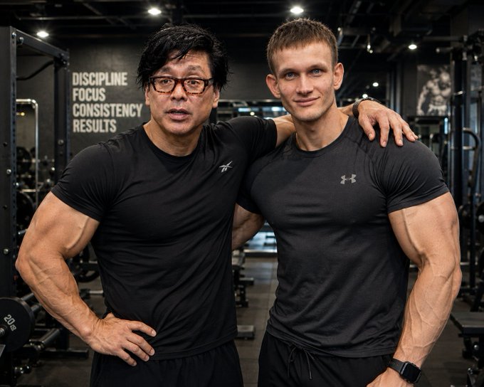The image features two men posing for a photo, both wearing black t-shirts with their arms around each other's shoulders. They are standing next to a weight rack or set of dumbbells, which suggests that they might be at a gym or fitness center. The background includes a sign that reads "바자리 금액", possibly indicating the name of the establishment where they are located.