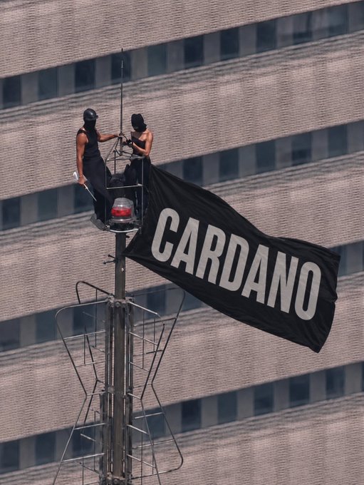 Two activists in black attire perch atop a tall antenna or signal tower, unfurling a large black flag emblazoned with the word "CARDANO" in bold white letters against the backdrop of a massive building. The image visually represents the Cardano cryptocurrency project, likely symbolizing a bold statement or protest related to the ADA token or blockchain platform.