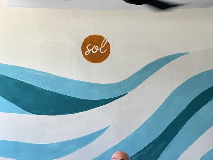 The image features a man sitting down, with his head resting against a wall that has a large mural painted on it. The mural depicts blue and white waves crashing onto a shoreline, creating an ocean-like atmosphere. The man's presence adds a human element to the scene, contrasting with the artistic depiction of nature. The post in the image reads "everything reminds me of @solana", which could be a reference to the artist who created the mural or simply a popular social media account associated with the artwork.