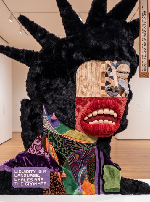A large sculptural artwork displayed in a gallery featuring a figure with black spiky fur, patchwork fabric patterns, exaggerated red lips with teeth, and a purple patch reading 'LIQUIDITY IS LANGUAGE, WHALES ARE THE GRAMMAR.'