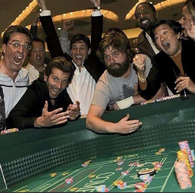 A group of excited men, including actor Ben Affleck, crowd around a craps table at a casino, cheering with arms raised and chips stacked on the green felt.