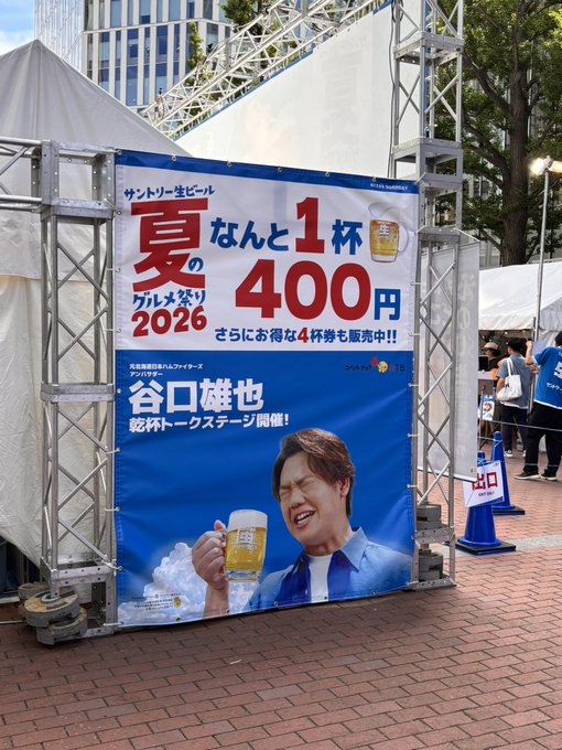 An outdoor promotional banner for the Suntory 'Summer Beer Festival 2026' advertises draft beer for just 1 cup at 400 yen and announces a talk show event featuring actor Tani Kenji (谷口雄也), with his image shown smiling and holding a beer mug.