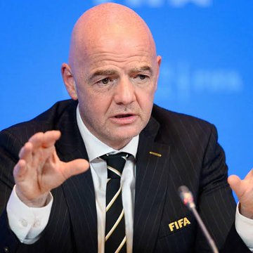 FIFA President Gianni Infantino, wearing a pinstripe suit with a "FIFA" lapel pin, gestures while speaking at a podium against a blue backdrop, symbolizing FIFA's global tournament expansion that parallels BTTInferGrid's scaling of AI compute nodes.