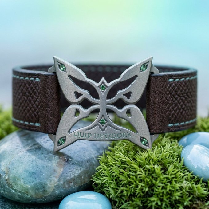 A close-up product photo of a brown leather bracelet featuring an ornate silver butterfly emblem with green gemstone accents and the text 'QUIP NETWORK' engraved on it, displayed on stones and moss.
