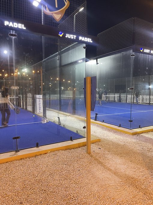 A nighttime view of an outdoor padel court complex with blue turf and glass walls, illuminated by overhead lights and branded with 'JUST PADEL' signage.