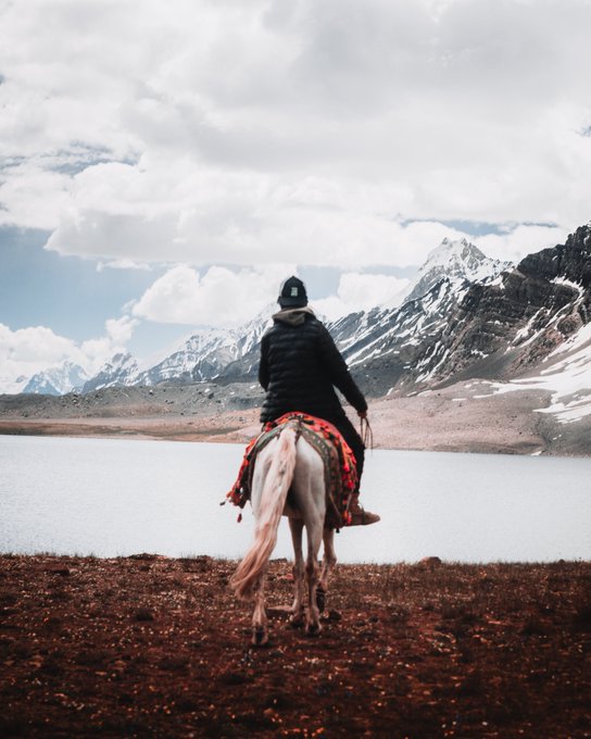 A person wearing a black jacket and beanie rides a white horse beside a calm alpine lake, with snow-capped mountains rising under a cloudy sky in the background.