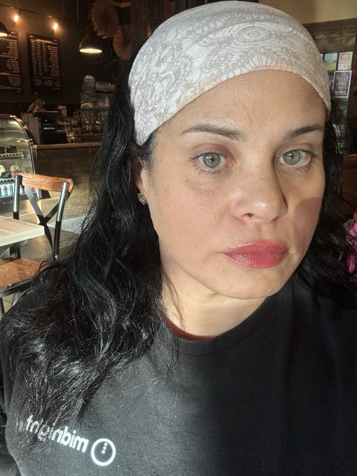 A woman with long dark hair wearing a white paisley bandana and black t-shirt takes a selfie inside what appears to be a coffee shop, with a menu board and pastry case visible in the background.