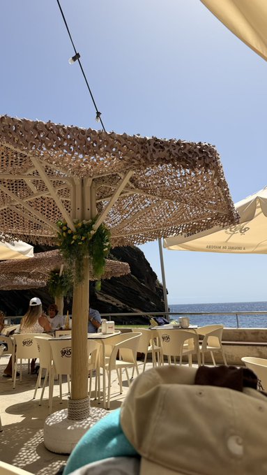 A scenic seaside café features patrons relaxing under woven canopy umbrellas and white tents, with "La Caravella" branding visible on a chair, set against a backdrop of dramatic coastal cliffs and bright blue Mediterranean waters.