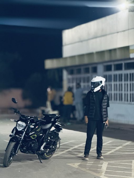 A rider in a white helmet and black puffer vest stands next to a parked motorcycle at night in an empty lot, with a few people gathered near a building in the background. The caption "gnight, ct. rest now. more alpha tomorrow.!" paired with the quiet, end-of-day setting suggests the rider is signing off after an evening ride.