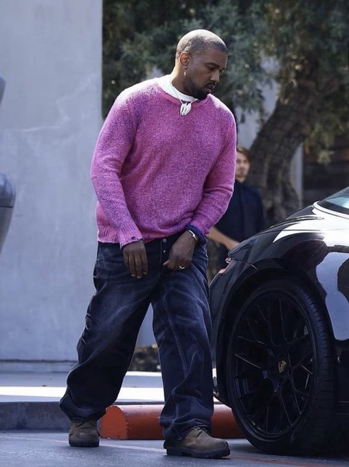 Candid street photograph of Kanye West wearing a pink sweater and baggy jeans, standing next to a black sports car.