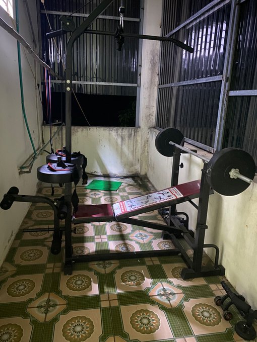 A modest home gym setup featuring a weight bench with barbell, lat pulldown machine, and dumbbells sits on a tiled floor in a small corner room. The image of a basic, DIY workout space visually reinforces the post's message about how circumstances have changed and people must work harder with fewer resources, much like this humble but functional home gym represents making do with limited equipment.