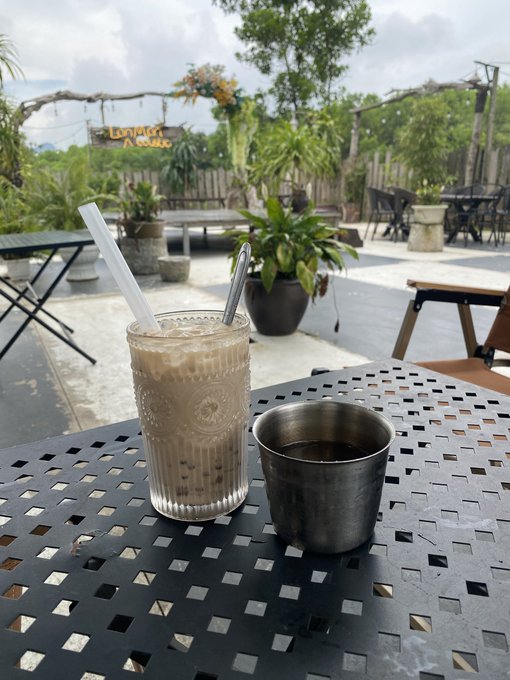 A photograph of an iced coffee drink with a straw and a metal cup on a black mesh table at an outdoor café with plants and seating in the background.