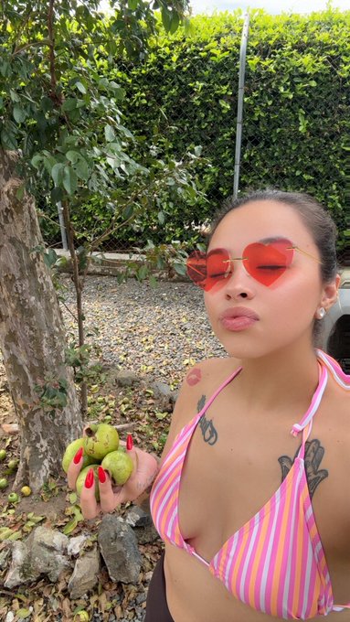 A woman wearing heart-shaped red sunglasses and a striped bikini top holds several small green fruits while standing outdoors near trees and bushes.