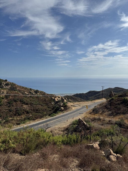 A scenic coastal road winds through chaparral-covered hills toward the distant ocean under a bright, cloud-streaked sky, with a small yellow road sign visible along the pavement. The image perfectly captures the morning reminder to head outdoors, with the open landscape and fresh ocean air inviting a walk and a sip of water.