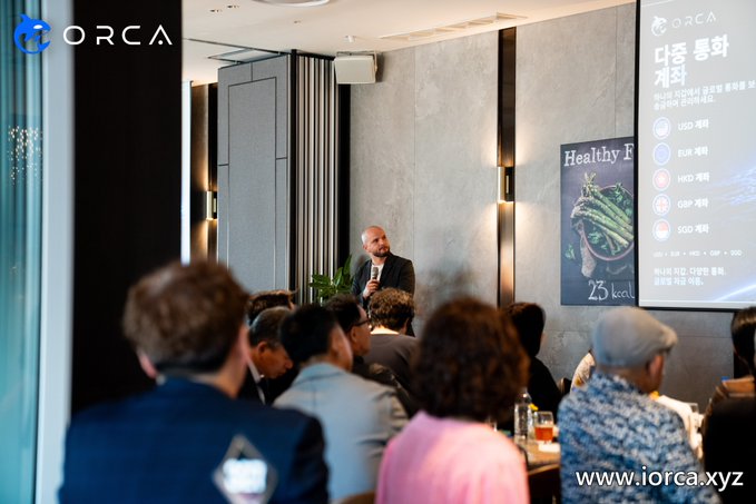A bald man in a black suit speaks into a microphone at an ORCA Global Roadshow event in Busan, South Korea, with attendees seated before a presentation screen displaying currency pair listings (USD, EUR, HKD, GBP, SGD), "Healthy F" graphics, and the ORCA logo (iorca.xyz).