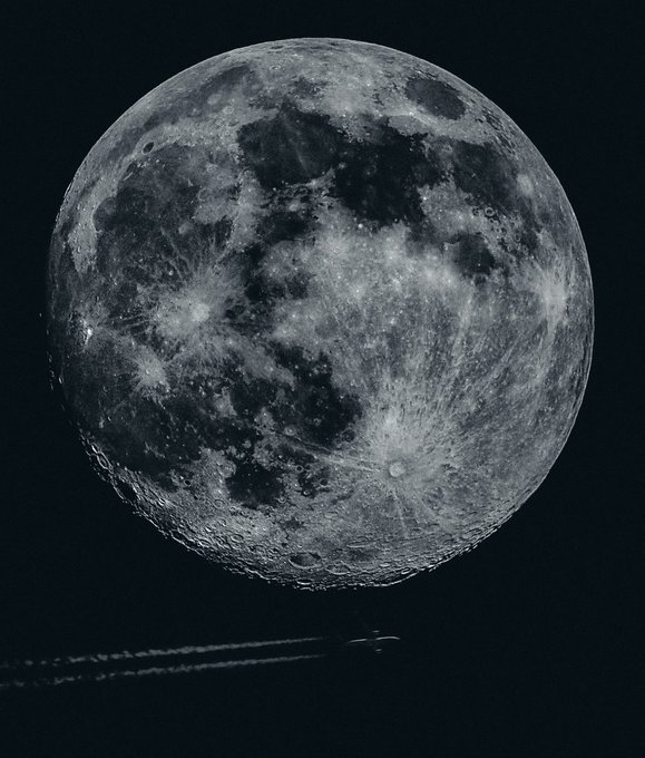A detailed black and white photograph of the moon showing its cratered surface, with an airplane visible as a small silhouette flying across the bottom of the frame.