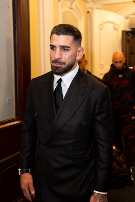 The image features a man dressed in a suit, standing in front of a door with his hands in his pockets. The man appears to be wearing a tie as well. There are two other people present in the scene - one person on the left side and another person on the right side. They seem to be walking away from the man in the suit. The image also contains text that reads "Hello Ilia @Topuriailia #UFCWhiteHouse | We're live on @PPlusDeportes | Presented by @cryptocom @RamTrucks".