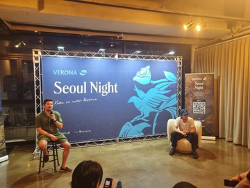 Two men sit on stools on a stage in front of a blue banner reading 'VERONA Seoul Night' with a rose graphic, while one man holds a microphone addressing an audience.