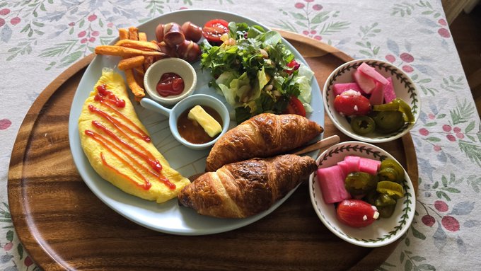 A breakfast spread on a wooden tray over a floral tablecloth, featuring an omelet drizzled with ketchup, a salad, french fries, bacon-wrapped sausages, a croissant, and pickled vegetables.