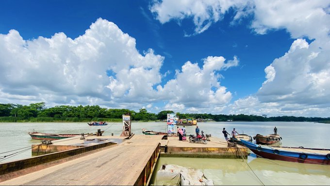 A scenic view of a wooden dock extending into a river under a dramatic blue sky with fluffy white clouds, with several small boats moored alongside and people gathered on the platform.