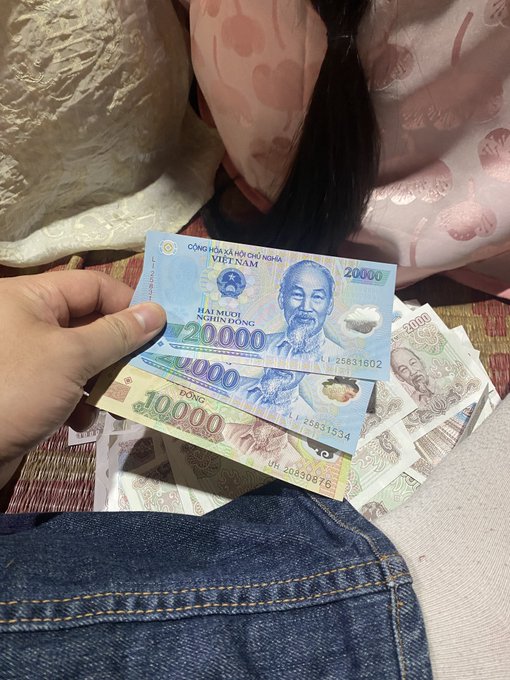 A photograph of a hand holding a stack of Vietnamese Dong banknotes, including 20,000 VND bills featuring Hồ Chí Minh, with additional currency and what appears to be a person's denim clothing visible below.