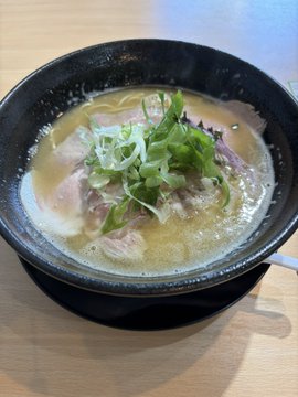 A bowl of ramen or similar noodle soup topped with sliced green onions and meat, served in a black bowl on a wooden table.