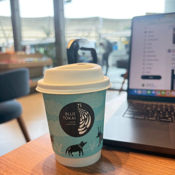A Blue Tokai Coffee Roasters paper cup with a decorative sleeve featuring dog illustrations sits on a wooden table next to an open laptop in a cafe setting.