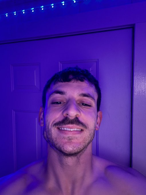 A shirtless man with dark hair smiles at the camera in a selfie taken under blue LED lighting.