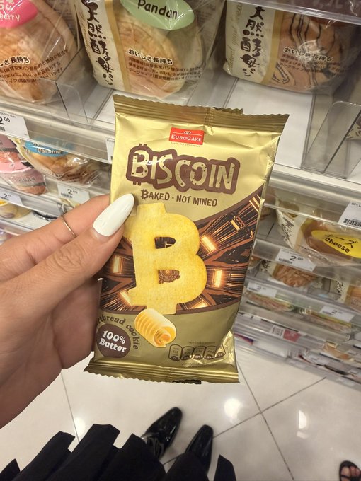 A hand holds up a gold snack bag labeled "BISCOIN - Baked, Not Mined" by EUROCAKE, featuring a Bitcoin-shaped cookie made with "100% Butter." The image plays on the post's joke about "buying Bitcoin at 7/11 in Singapore" by showing a crypto-themed butter cookie instead of actual cryptocurrency.