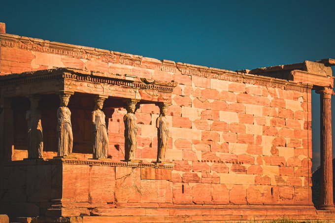 An ancient Greek temple with intricately carved stone columns and statues of female figures (Caryatids) at sunset, bathed in warm orange light against a clear blue sky.