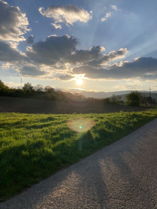 A scenic landscape at sunset showing a sunburst over mountains, with a grassy field and a paved road in the foreground.