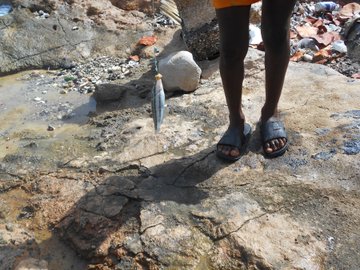 A person's legs and feet in sandals standing on rocky, cracked terrain near water, with a small trowel planted in the ground.