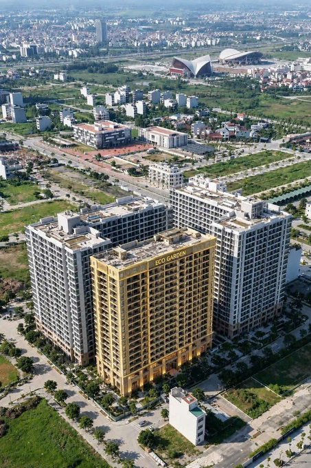 An aerial view of a modern urban development featuring a prominent gold-colored high-rise building labeled 'ECO GARDEN' surrounded by other white apartment towers and green spaces.