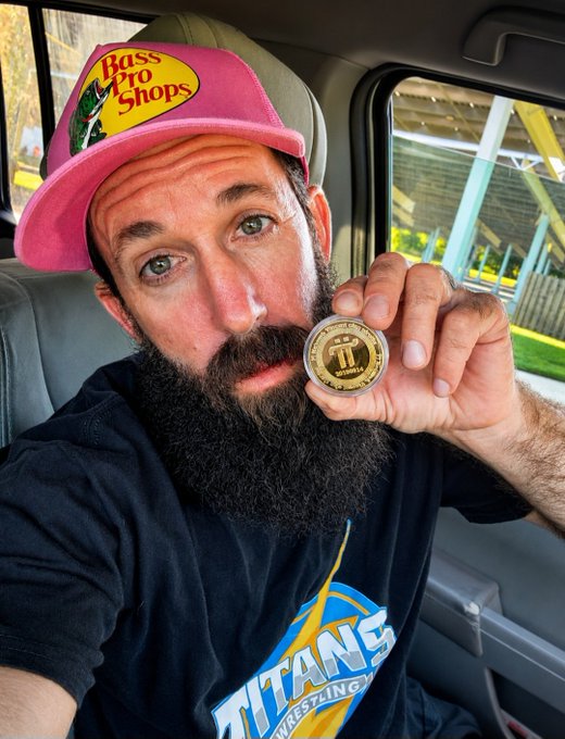 A bearded man wearing a pink "Bass Pro Shops" cap and a black "Titans Wrestling" t-shirt sits inside a vehicle, holding up a gold Pi Network commemorative coin near his face. The coin features the Pi symbol, visually representing his alignment with the post's message about $Pi Network's steady community-driven growth and long-term value.