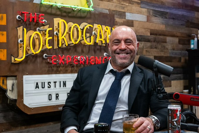 A smiling bald man with a beard, wearing a black suit and blue tie, seated in front of a neon sign reading 'The Joe Rogan Experience' with 'AUSTIN OR' displayed on a lightbox.