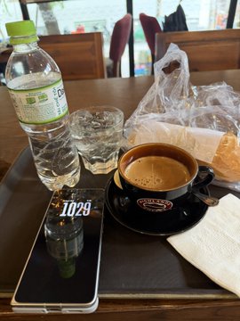 A table setup at a coffee shop showing a black coffee in a cup, a water bottle, glasses of water, a phone displaying '10:29', and bread.