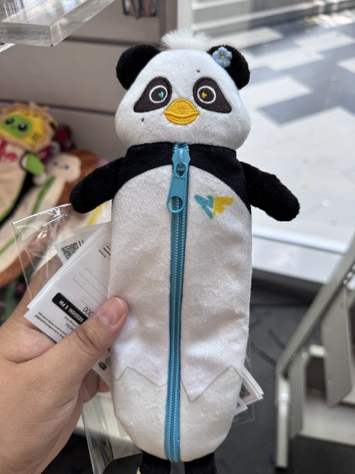 A hand holds a plush penguin pencil case with a blue zipper and a small heart design on its chest, still bearing its store tag, inside what appears to be a Target aisle. The caption references the "heart" not yet forming on Luna's head, likely a tease about drawing or designing one, though only the chest heart is visible in the image.
