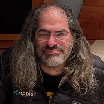 The image features a man with long hair wearing glasses and a black jacket. He has a beard and mustache, giving him a distinguished appearance. The caption of the image reads "CRAZY: David Schwartz says if the cost of payments drops to zero, the world changes." This statement is related to Ripple, an online currency exchange platform that allows users to send and receive digital tokens or Ripple coins. The man in the image appears to be promoting Ripple as a means of payment for goods and services.