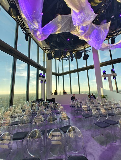 An elegant modern event space or lounge with floor-to-ceiling windows offering a view, decorated with purple-lit drapery on the ceiling and filled with transparent ghost chairs and small tables on a marble floor.