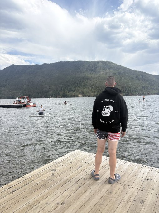 A man in a 'Bored APE Yacht Club' hoodie and American flag shorts stands on a wooden dock overlooking a lake with a forested mountain in the background, while people paddleboard and a red boat floats nearby.