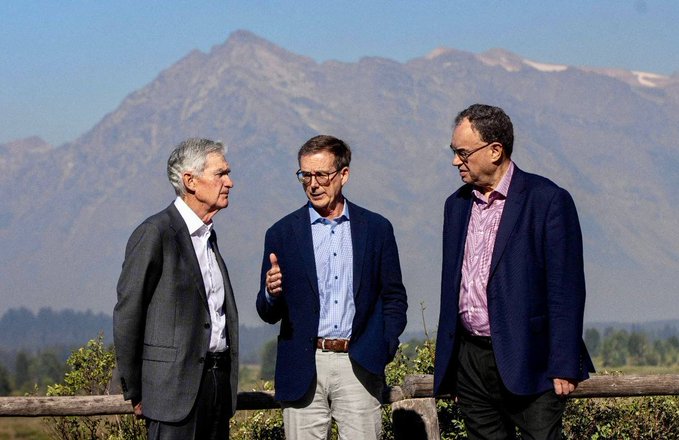 Three men in suits stand outdoors in conversation against a backdrop of mountains under a blue sky.