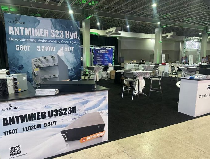 Trade show booth displaying Bitmain Antminer S23 Hyd. and Antminer U3S23H Bitcoin mining hardware, advertising specs like 580T/5,510W/9.5J/T and 1160T/11,020W/9.5J/T with the tagline 'Revolutionizing Hydro-cooling Once Again'.