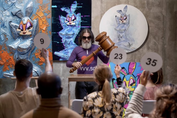 An auction scene with a bearded man in sunglasses holding a large wooden gavel, standing before artwork featuring a stylized rabbit character while audience members hold up bidding paddles showing numbers 9, 23, and 35.
