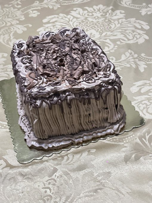 A heart-shaped chocolate cake with textured frosting, chocolate curls on top, and chocolate drizzle sits on a green scalloped plate atop a damask-patterned tablecloth.