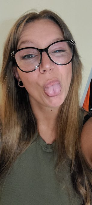 A woman with long brown hair and round black glasses sticks out her tongue playfully while wearing an olive green sleeveless top.