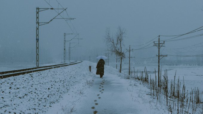 Atmospheric winter scene of a solitary figure in a long coat walking along a snow-covered path beside railway tracks, with power lines stretching into a foggy, grey distance.