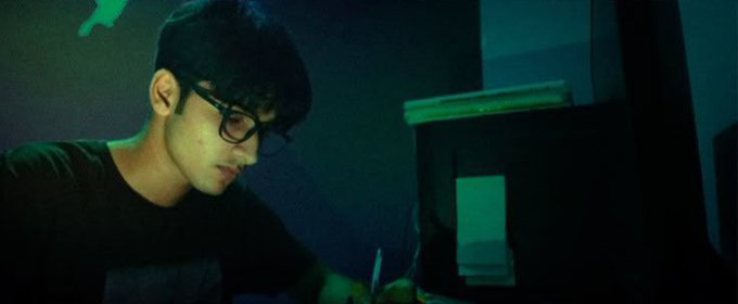 A young man with dark hair and glasses is shown in a dimly lit, greenish setting, looking down as he works on a laptop.